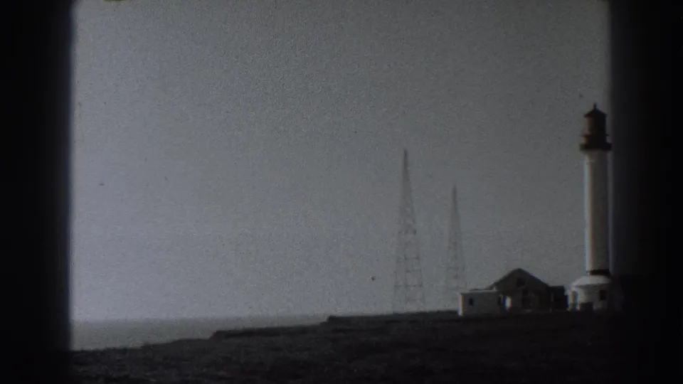Fort Ross area view with a tall coastal tower, radio masts, low buildings, open ground, and the soft contrast of 1938 black-and-white 8mm film.