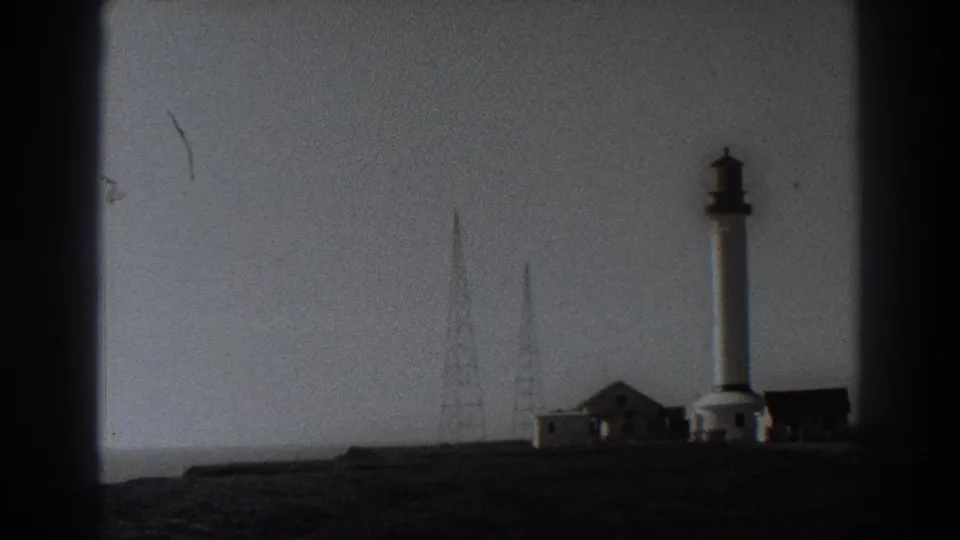 Black-and-white Fort Ross home-movie frame showing a tower, radio masts, small buildings, and open coastal terrain in soft 8mm texture.