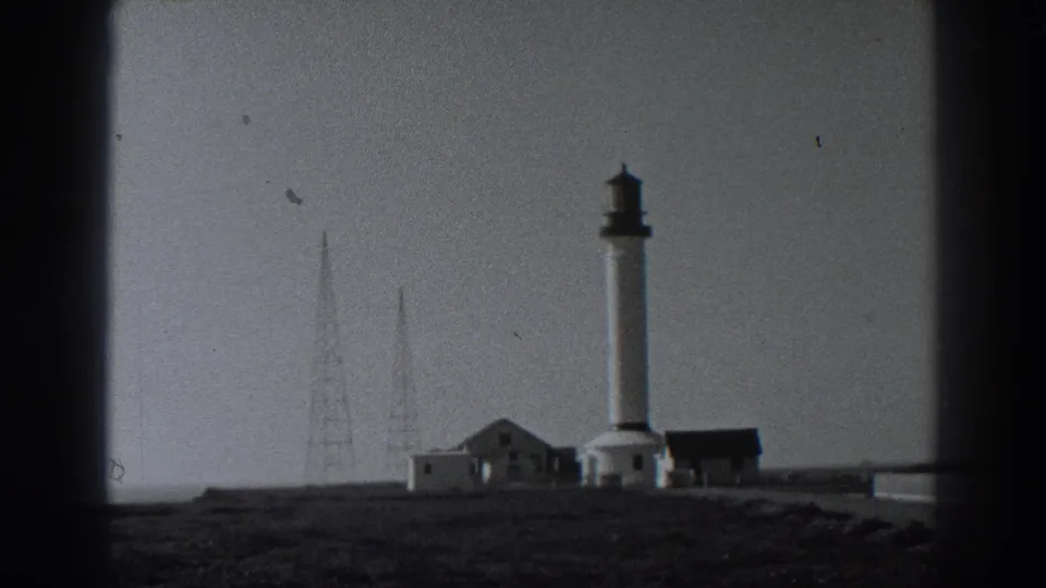 1938 Fort Ross home-movie frame showing a lighthouse, outbuildings, radio masts, and grassy coastal ground.