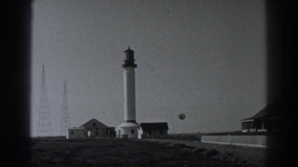 1938 Fort Ross frame showing the lighthouse complex, radio masts, grassy headland, and low wall in front.
