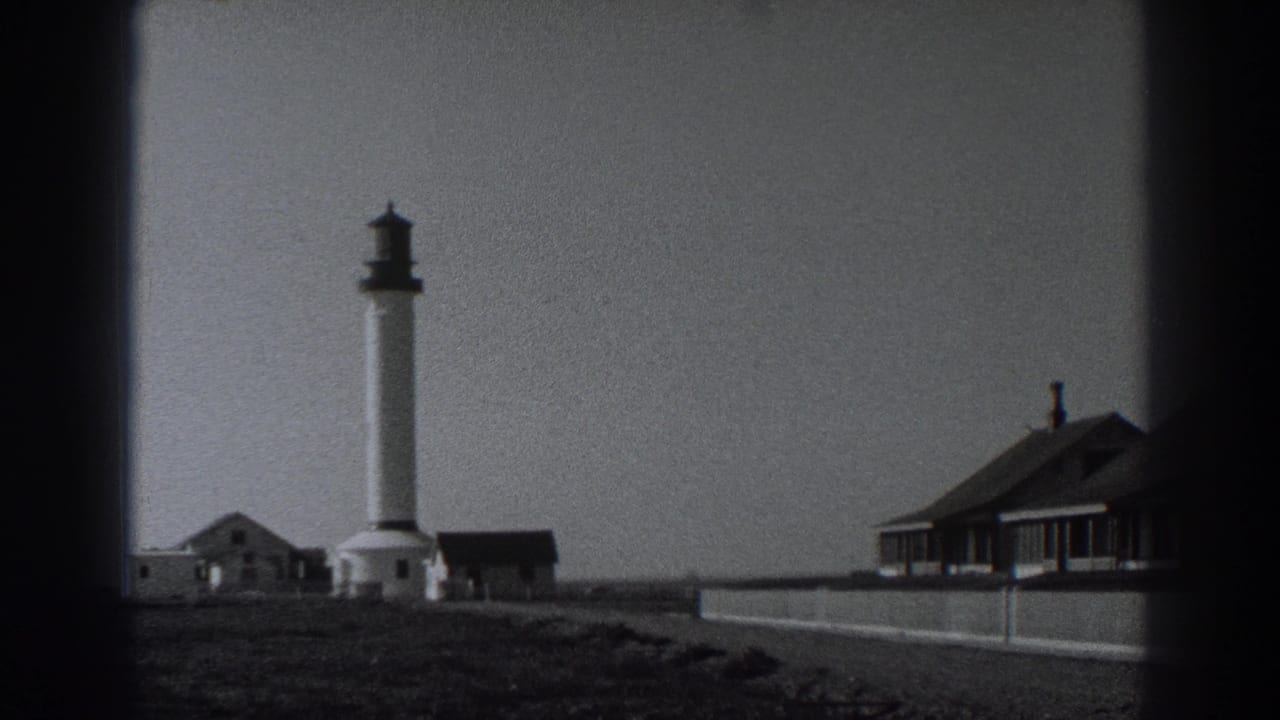 White lighthouse and small coastal buildings beside a low wall under open sky.