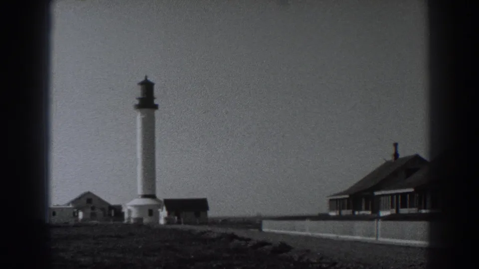 1938 Fort Ross coastal view with a white lighthouse, small outbuildings, rough shoreline ground, and cottages at the right edge.