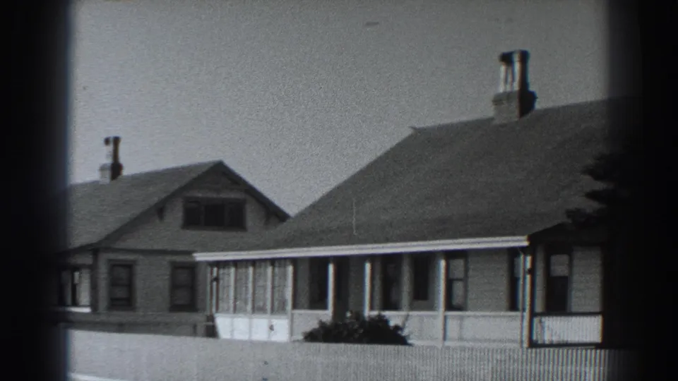 Closer 1938 Fort Ross view of a gabled house with chimney, porch windows, pale fence, and low shrubs in front.
