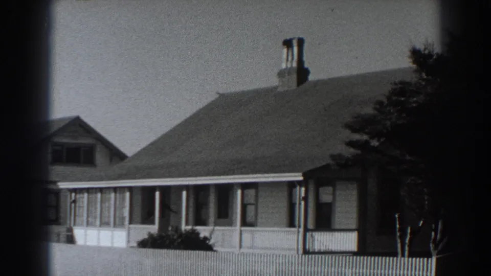 Fort Ross house exterior in 1938 with a broad roof, chimney, porch windows, front fencing, shrubs, and a tree along the edge.
