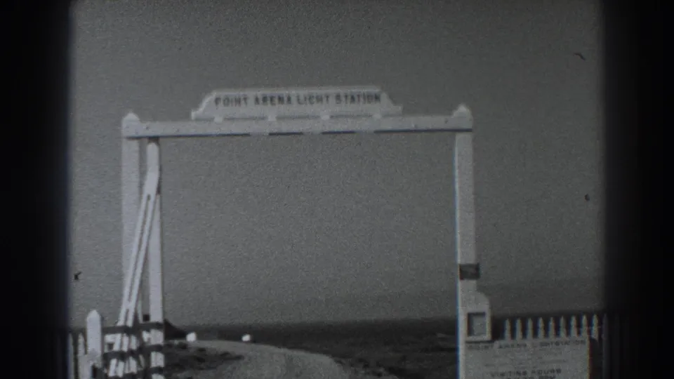 Black-and-white 8mm view of the Point Arena Light Station sign structure with fence posts and open coastal terrain beyond.