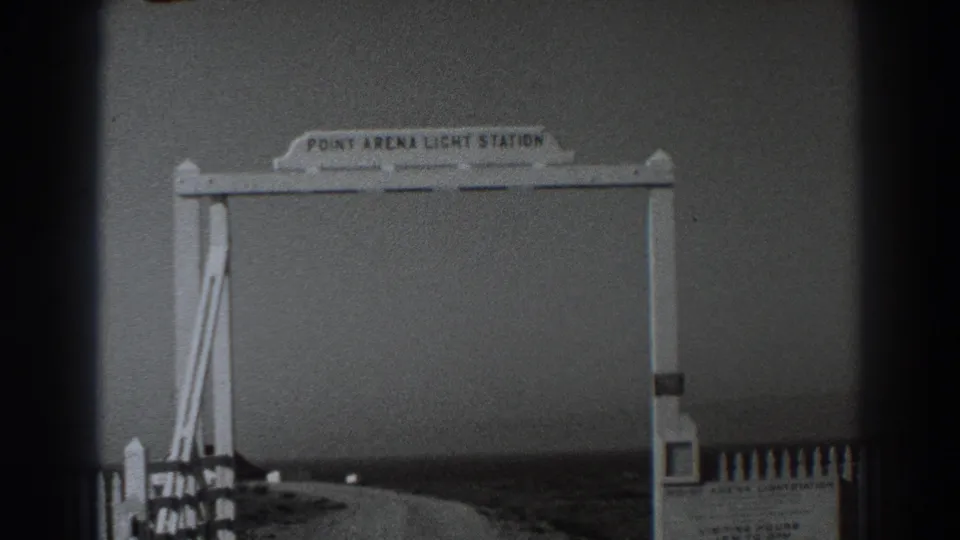 1938 home-movie frame of the Point Arena Light Station arch with a curving road, roadside posts, and soft coastal background.