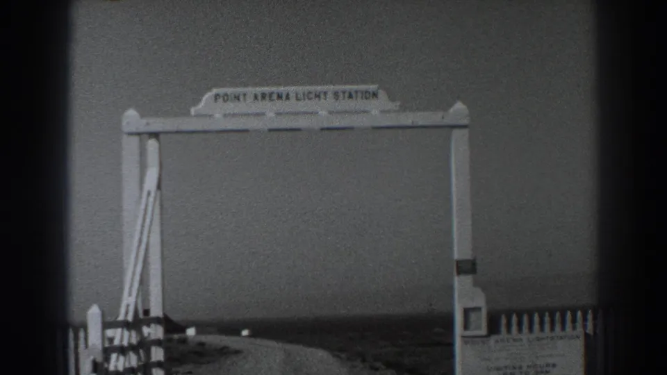 Black-and-white 8mm frame showing the Point Arena Light Station entrance arch, fence posts, and coastal roadside view in California.