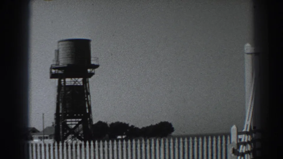 1938 California home-movie frame of a dark water tower, picket fence, low trees, and part of the light station entrance structure.