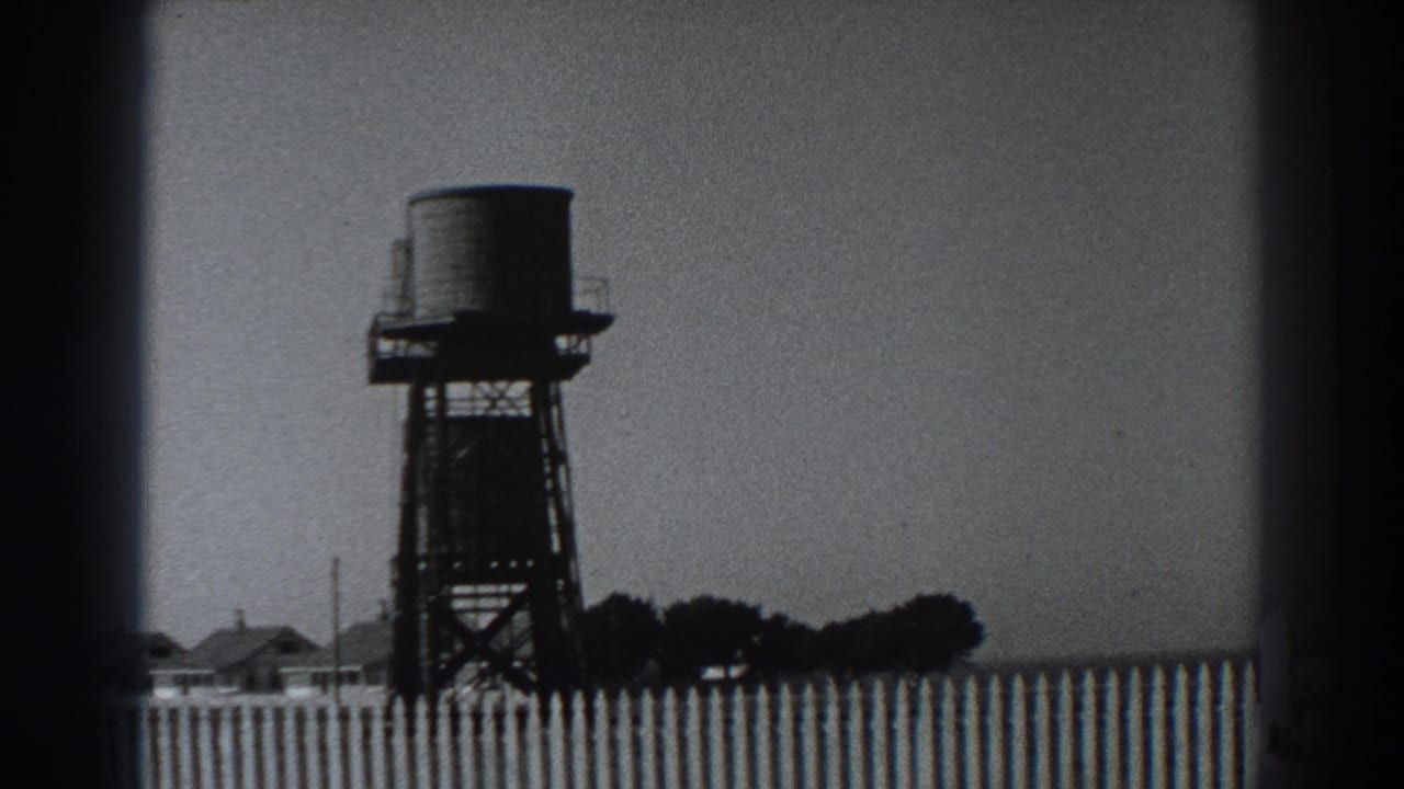 1938 Fort Ross-area home-movie frame showing a raised water tank, fence, low buildings, and distant trees under a pale sky.