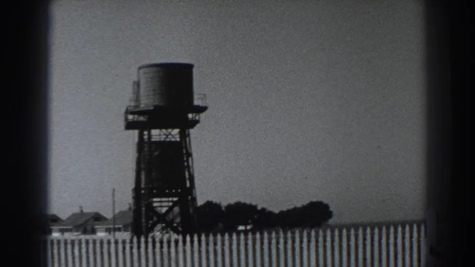 1938 Fort Ross-area home-movie frame showing a raised water tank, fence, low buildings, and distant trees under a pale sky.