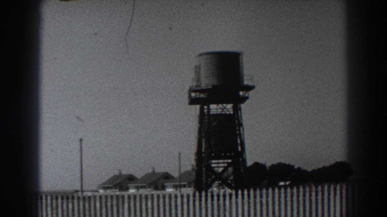 Fort Ross, California 1938 frame with a water tank tower above a fence, low rural buildings, utility poles, and a tree line.