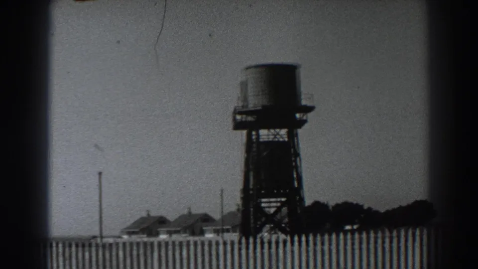 Fort Ross, California 1938 frame with a water tank tower above a fence, low rural buildings, utility poles, and a tree line.