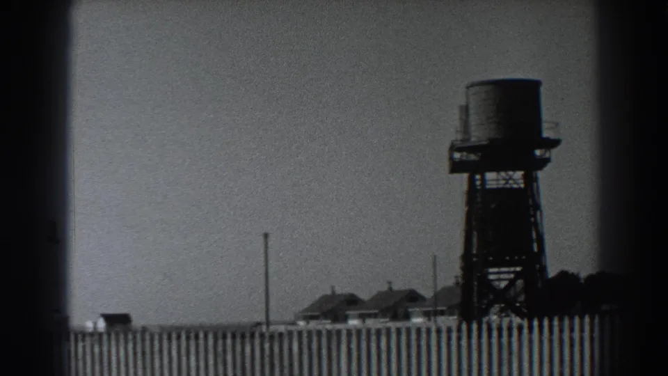 1938 rural Fort Ross home-movie view with the water tank shifted right, a picket fence, small buildings, poles, and dark trees.
