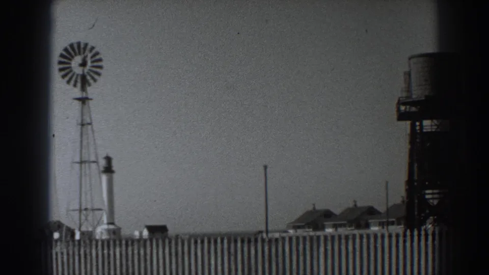 Fort Ross-area 1938 frame showing a windmill, partial water tank tower, fence, low buildings, poles, and broad pale sky.