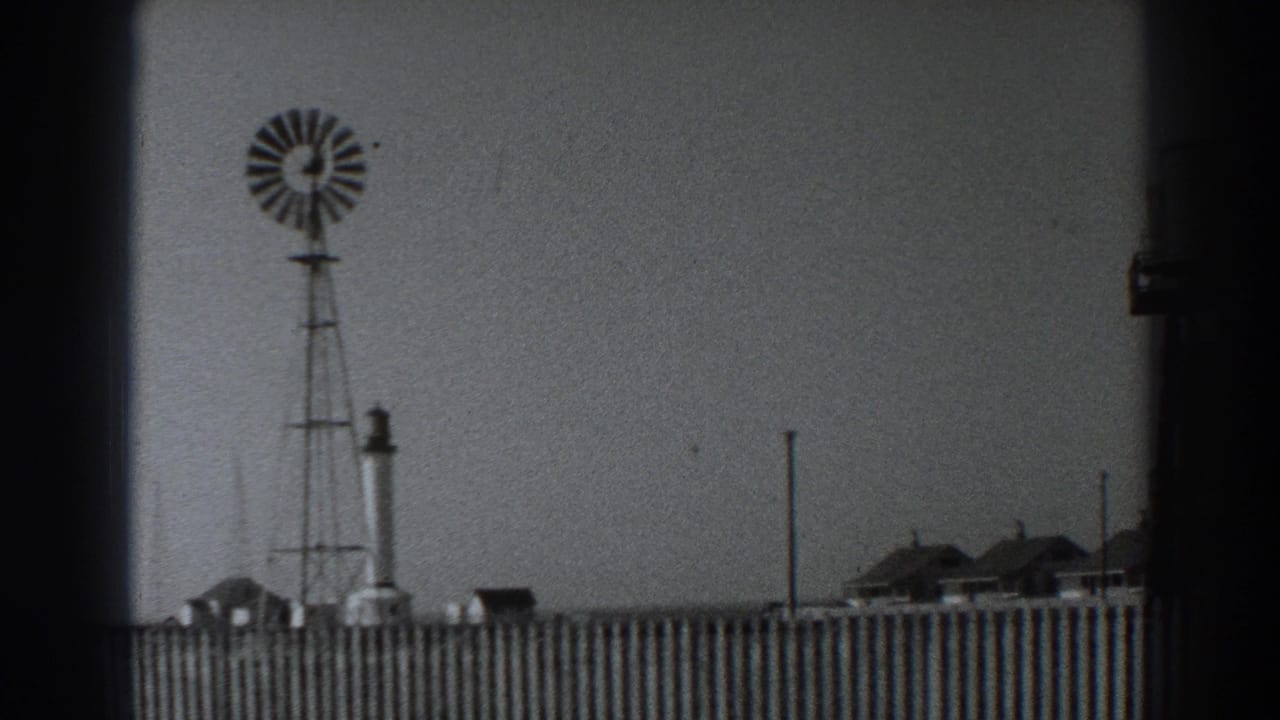 1938 Fort Ross view of a windmill above a wooden fence, with a tower and low buildings under a pale sky.