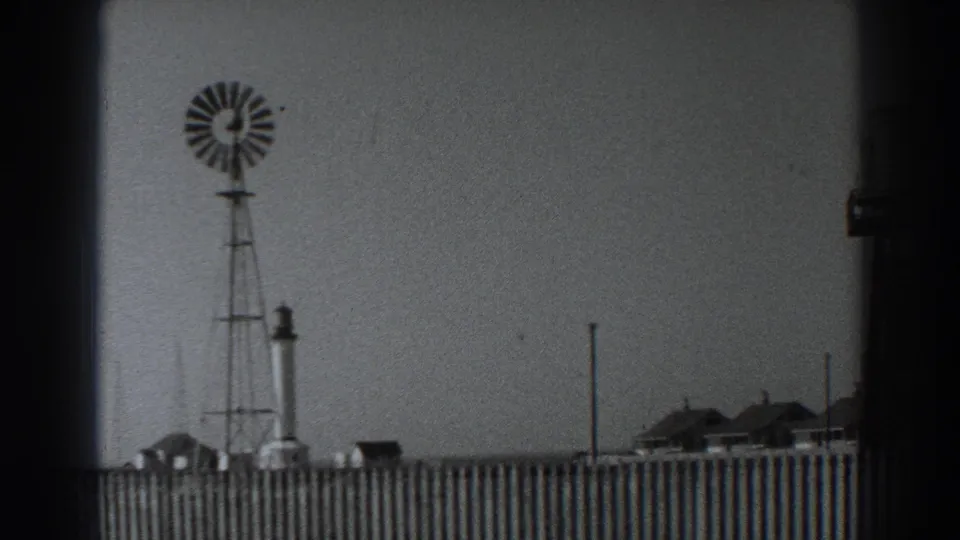 1938 Fort Ross view of a windmill above a wooden fence, with a tower and low buildings under a pale sky.
