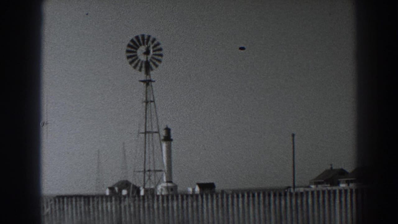 Fort Ross windmill and tower seen past a wooden fence, with small buildings along the horizon in 1938 home-movie footage.