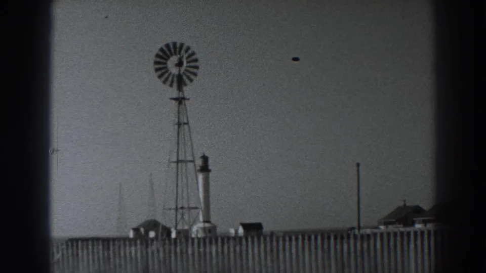 Fort Ross windmill and tower seen past a wooden fence, with small buildings along the horizon in 1938 home-movie footage.