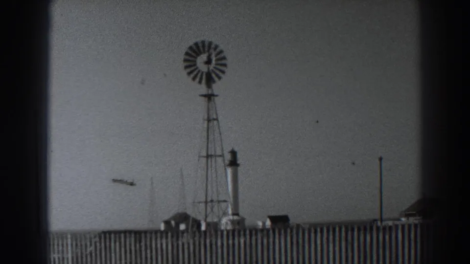 Windmill framed above a wooden fence at Fort Ross, with a tower, poles, and small structures visible in the 1938 scene.