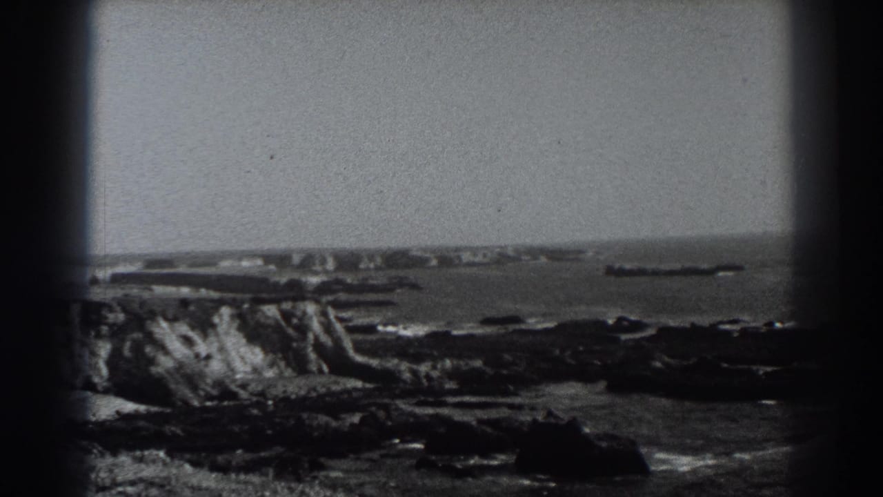 Rocky coast and ocean horizon with a long dark vessel-like shape offshore.