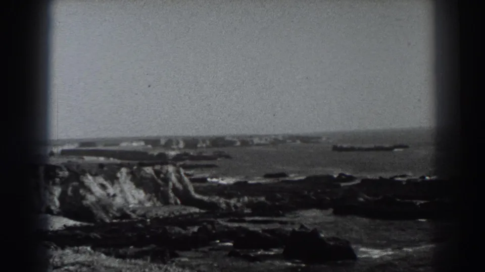 Fort Ross-area coastal frame with cliffs, dark shoreline rocks, open water, and a distant vessel-like form on the horizon.