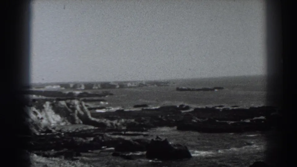 1938 8mm coastal view near Fort Ross with low rocky bluffs, surf, sea, and soft black-and-white film texture.