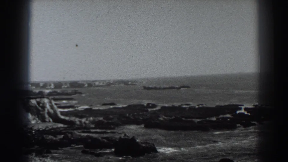 Fort Ross-area shoreline frame showing rocks, breaking waves, open water, and a distant dark offshore shape in soft focus.