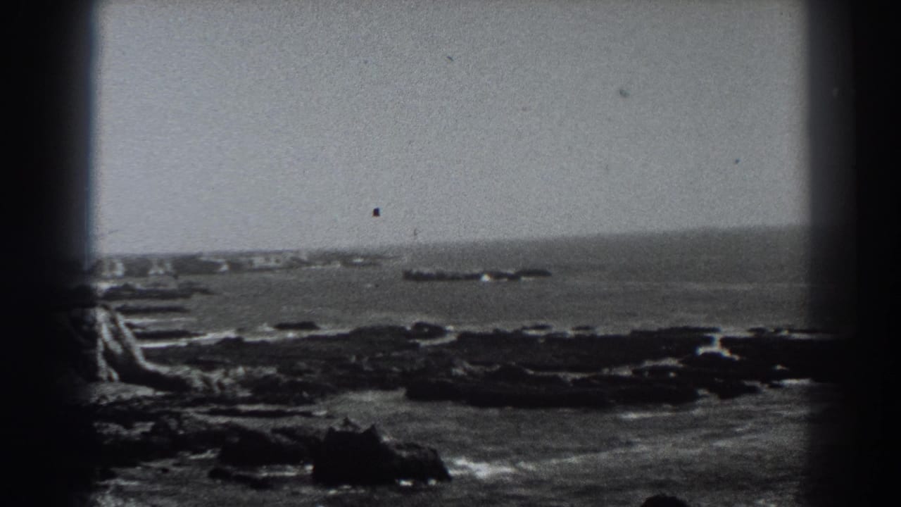 1938 Fort Ross, California 8mm frame showing a rocky nearshore, breaking surf, open water, and a distant low offshore rock formation within dark film-frame borders.