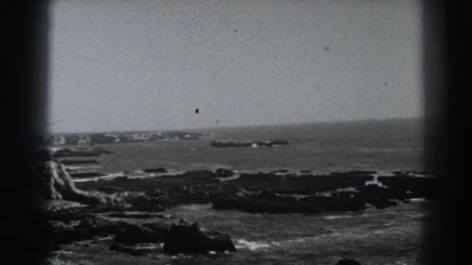 1938 Fort Ross, California 8mm frame showing a rocky nearshore, breaking surf, open water, and a distant low offshore rock formation within dark film-frame borders.