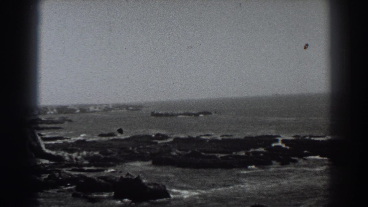 1938 Fort Ross, California 8mm frame holding on a wide rocky coastal view with white surf around dark rocks and a low offshore formation near the horizon.