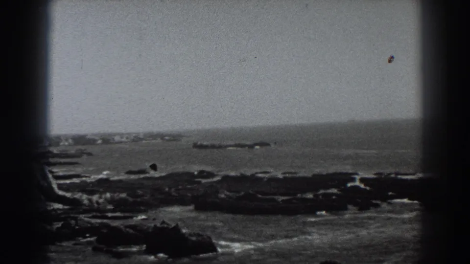 1938 Fort Ross, California 8mm frame holding on a wide rocky coastal view with white surf around dark rocks and a low offshore formation near the horizon.