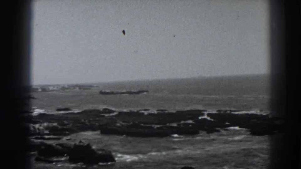 1938 Fort Ross, California 8mm frame showing rough water moving through a dark rock field, with a distant offshore rock formation and soft film grain across the sky.