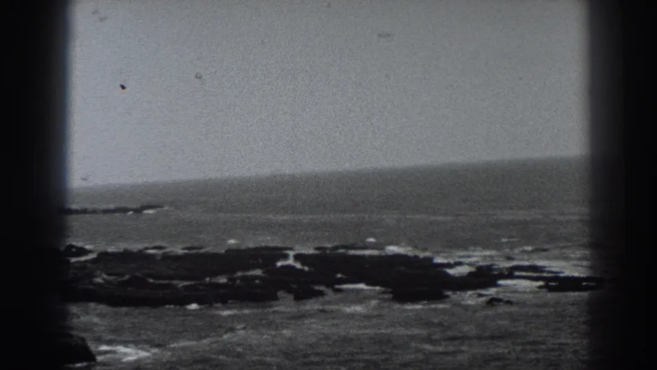 Black-and-white 8mm view of the Fort Ross coast, with low rocks, surf, and a hazy Pacific horizon.