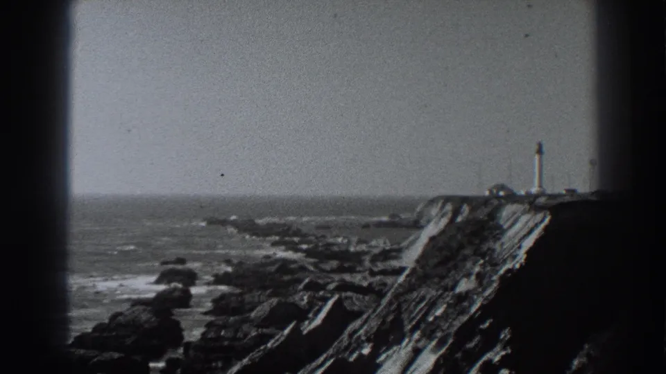 Black-and-white 8mm view of the Fort Ross shoreline, with surf, dark rocks, a cliff, and a distant lighthouse-like tower.
