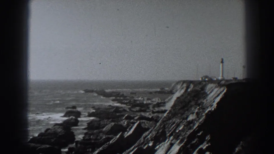 1938 Fort Ross, California home-movie frame showing a lighthouse on a rugged cliff above surf and rocks under an overcast sky.