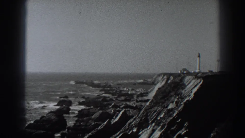 Black-and-white 8mm frame of the Fort Ross shoreline, with waves, rocks, cliff face, and a distant lighthouse near the horizon.