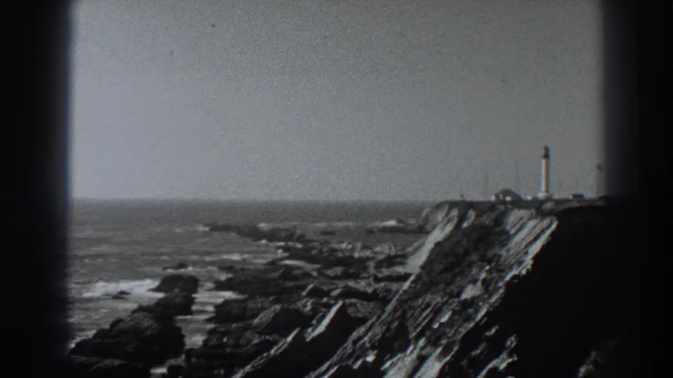 1938 Fort Ross, California home-movie frame showing a distant lighthouse-like tower on a rugged cliff above surf and rocks.