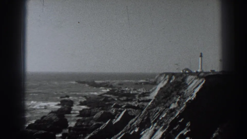 Fort Ross coastal view on 8mm film, with a lighthouse-like tower at right and choppy ocean beyond the rocks.