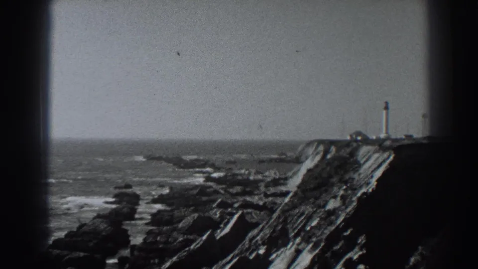A grainy 1938 Fort Ross frame looks across rocks and surf toward a clifftop lighthouse-like tower in the distance.