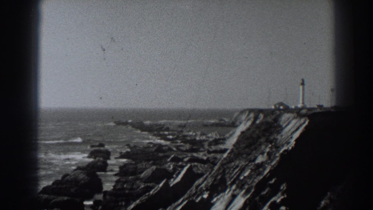 Distant lighthouse above rugged rocks and ocean water.