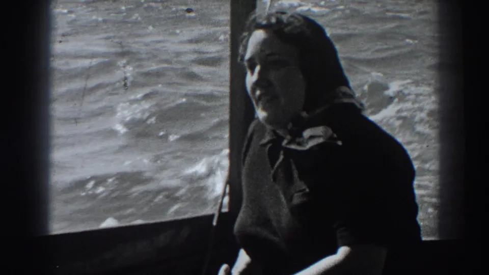 A seated woman in dark clothing turns toward the camera within the boat, with sunlit water visible through the window behind her.