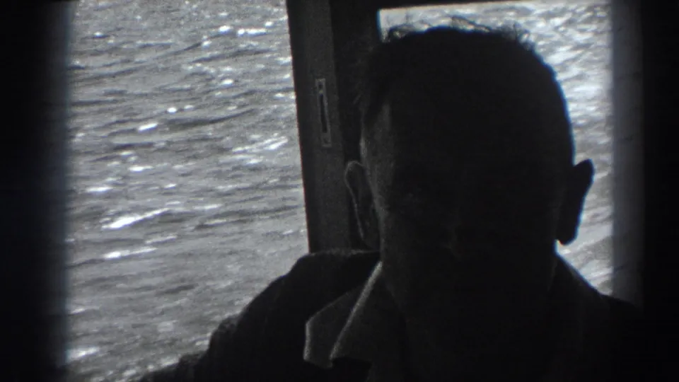 A balding man faces forward inside the boat cabin, his features largely obscured by shadow as Fort Ross-area water glints through the window.