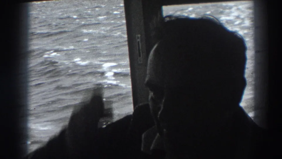 The passenger turns into profile and appears to raise a hand near his forehead, with bright rippling ocean visible through the boat window.