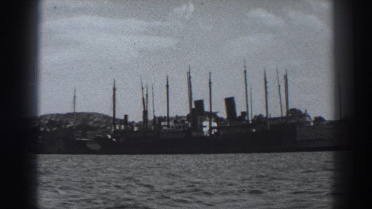 1938 Fort Ross 8mm frame showing a large docked ship in silhouette, rippled harbor water, shoreline, and cloudy sky.