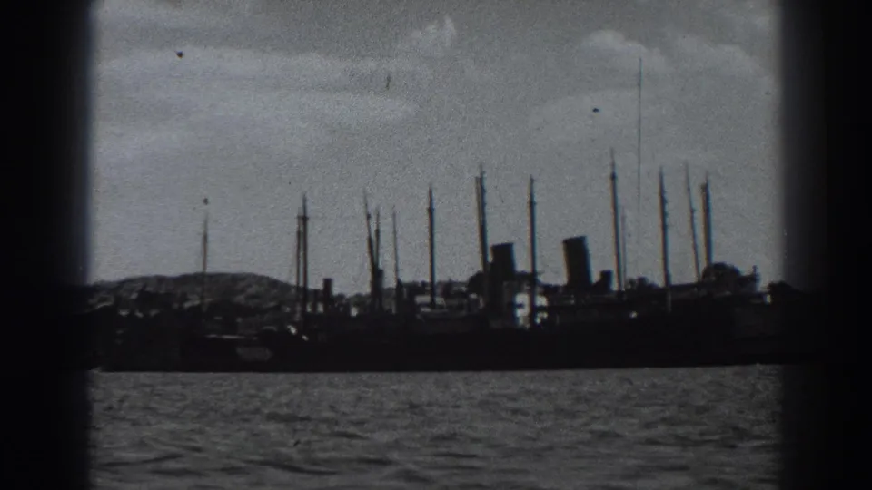 Fort Ross harbor view from 1938 8mm footage, with a silhouetted ship, tall masts, smokestack shapes, and water in front.