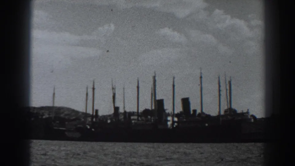 1938 8mm frame near Fort Ross showing distant ships and tall masts silhouetted across open water under clouds.