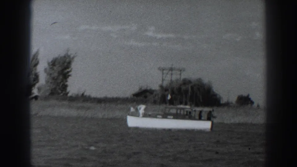 1938 Fort Ross home-movie frame showing a small white motorboat moving across water with trees, a low structure, and utility poles behind it.