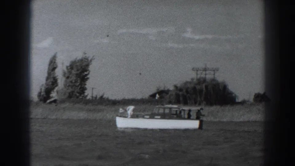 1938 Fort Ross frame with a small white motorboat gliding past a grassy shoreline, distant trees, and utility poles under a bright sky.