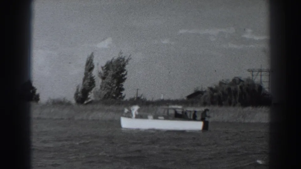 Fort Ross 1938 home-movie frame showing the motorboat farther across rippled water with trees, a low structure, and utility poles onshore.