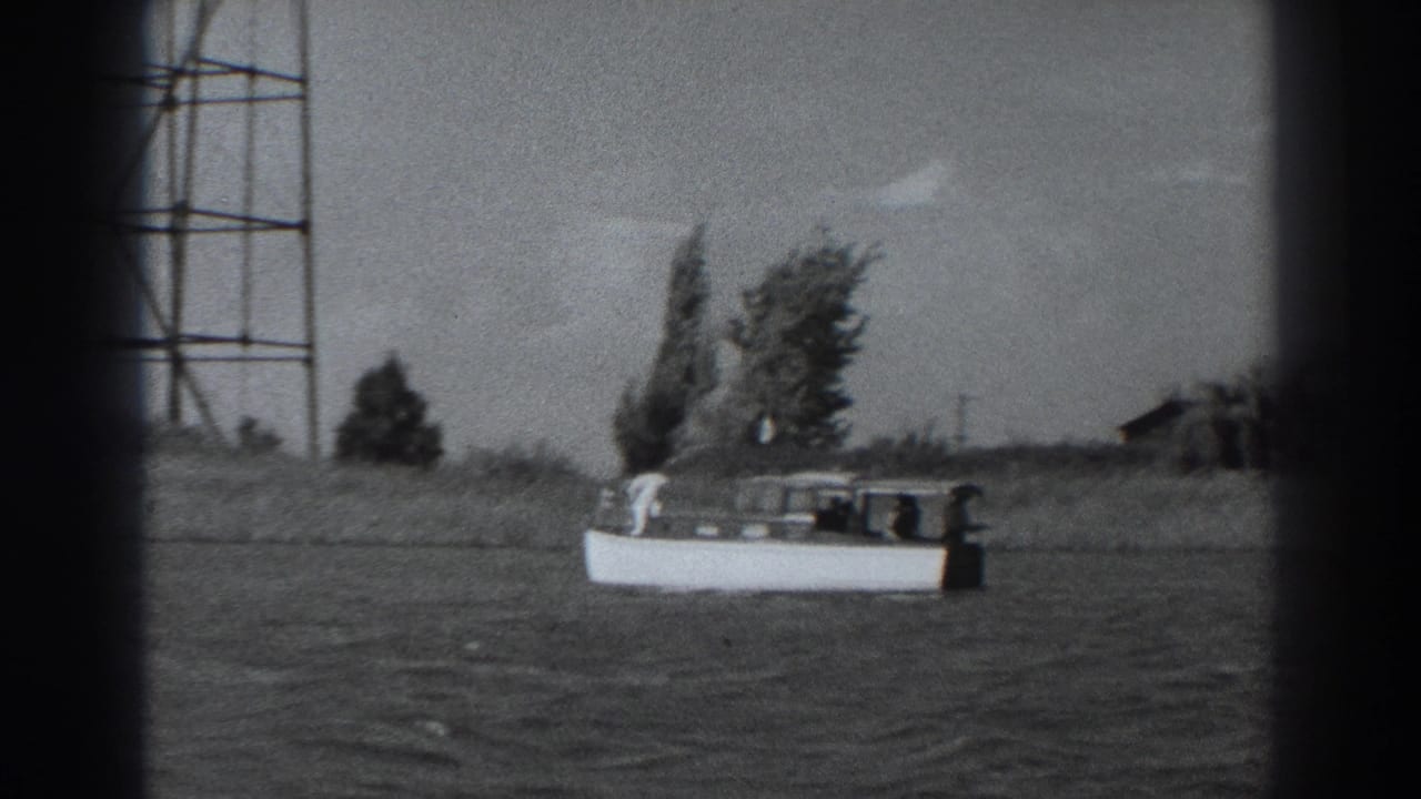 1938 Fort Ross home-movie frame showing a small white motorboat offshore, with trees and a tall metal tower along the background shoreline.